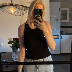 Svart ribbad ärmlös topp med öppen rygg - Säljer en svart ribbad topp från Mango med tight passform och öppen rygg. Toppen är ärmlös och har en snygg cut-out detalj bak som ger en unik look. Köpt i Spanien och är knappt använd, strl M🖤