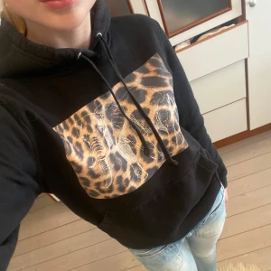 Svart hoodie med leopardtryck - Sjukt snygg svart hoodie med huva och snörning. På bröstet finns ett stort leopardmönstrat tryck med en vit elefantillustration ovanpå. Sjukt trendig och jättemjukt innertyg❤️