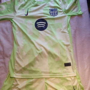 FC Barcelona bortaställ Nike ljusgrön - FC Barcelona bortaställ från Nike i ljusgrönt och vitt med geometriskt mönster. Tröjan har korta ärmar, rund hals, FCB-emblem och sponsorloggor på bröstet. Matchande shorts med resår i midjan och klubbmärke. Tillverkad i lätt och ventilerande polyester.