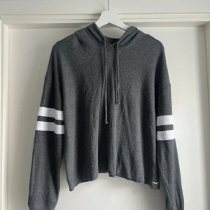 Grå hoodie från Hollister med ränder - Grå ribbad hoodie från Hollister Must-Have Collection. Croppad modell med huva och snörning, vita dubbla ränder på ärmarna och lång ärm. Liten broderad logga nertill. Perfekt för en chill och sportig look.