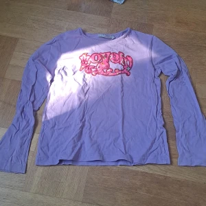 Lila långärmad tröja  - Lila långärmad tröja från FLY med coolt tryck på bröstet där det står 'Lovely' i rosa och blommigt mönster. Tröjan har rund halsringning och är gjord i mjuk bomull. Barnstorlek 146/152 men passar xs, eventuellt s.