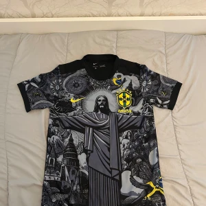 Nike Brasil Dri-FIT fotbollströja svart - Unik svart Brasilien fotbollströja från Nike med Dri-FIT-material. Framsidan har ett detaljerat konstnärligt tryck med Kristusstatyn, brasilianska symboler och gula detaljer. Korta ärmar med mönstrade partier och CBF-logga på bröstet. Perfekt för dig som vill sticka ut på planen.