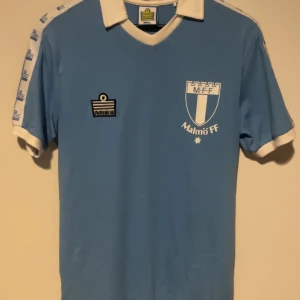Malmö FF retro fotbollströja Admiral 1979 - Klassisk ljusblå Malmö FF fotbollströja från Admiral med vit krage och vita detaljer på ärmarna. Stort klubbmärke på bröstet och Admiral-logga. Perfekt för dig som vill sticka ut på läktaren eller samla på retrotröjor. Använd enstaka gång och köpt i MFF shopen.