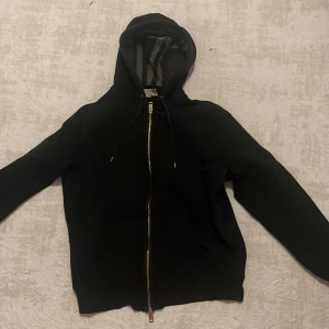 Svart hoodiejacka från Burberry M - Svart hoodiejacka från Burberry med dragkedja och klassisk rutig insida på huvan. Jackan har broderad logga på bröstet och snörning i huvan. Perfekt för dig som vill ha en stilren och lyxig look med streetvibe.