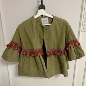 Grön overshirt med fransar från Zara - Säljer en grön overshirt från Zara med vida trekvartsärmar och snygga broderade band med röda och orange fransar. Jackan är i bomull och har tryckknappar framtill. Perfekt för dig som vill sticka ut med coola detaljer.