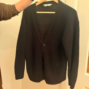 Svart stickad kofta med knappar - En svart stickad kofta från H&M med djup v-ringning och knappar framtill. Koftan har långa ärmar och ribbade muddar vid ärmslut och nederkant. Perfekt för lager-på-lager och chill vibes.