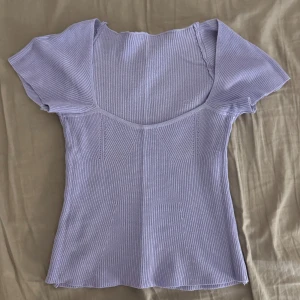 Ljuslila ribbad topp med kort ärm - Superfin ljuslila topp med ribbad struktur och korta ärmar. Toppen har en snygg v-ringning och är figurnära utan att vara för tight. Perfekt att styla med jeans eller kjol för en fräsch look.