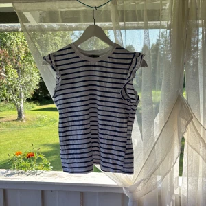 Randig volangtopp från SHEIN XS - Säljer en vit och marinblå randig topp från SHEIN i storlek XS. Toppen har volangdetaljer vid ärmöppningarna och är ärmlös med rund halsringning. Perfekt för en fräsch och somrig look. Materialet är mjuk bomull.