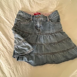 Jeanskjol med volanger från Lindex - Snygg jeanskjol från Lindex med volanger och klassiska fickor fram. Kjolen har en ljusblå tvätt och markerade sömmar som ger en cool retrovibe. Perfekt för dig som gillar denim och vill ha en avslappnad men trendig look.