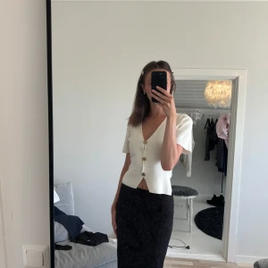 Vit kortärmad blus med knappar - Säljer en stilren vit blus med korta ärmar och v-ringning. Toppen har snygga guldiga knappar framtill och är lätt croppad för en modern look. Perfekt att matcha med både kjol och jeans för en clean och fräsch stil.