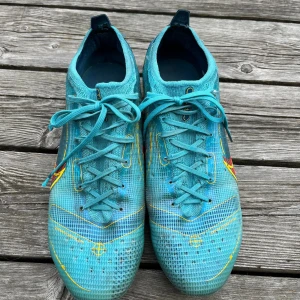 Turkosa Nike fotbollsskor med dobbar - Säljer ett par turkosa Nike fotbollsskor med snörning och coola gula och röda detaljer på sidorna. Skorna har rund tå och plastsula med blå och gula dobbar för bra grepp på planen. Ovandelen är i syntetmaterial med rutigt mönster och Nike-logga.