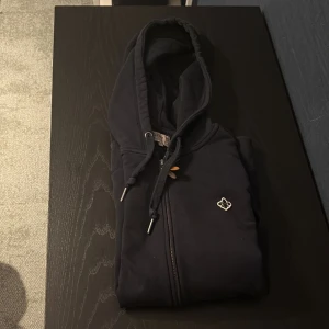 Mörk blå hoodie med dragkedja från Morris - Mörk blå hoodie från Morris i storlek M med dragkedja och huva. Tröjan har snörning i halsen, ribbade muddar och en broderad logga på bröstet. Perfekt för dig som gillar stilrena och klassiska plagg med snygga detaljer.