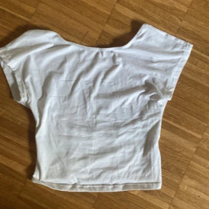 Vit croppad topp med öppen rygg - Säljer en vit croppad topp i bomull med korta ärmar och en snygg öppen rygg. Toppen har en enkel och clean look framtill och en djup, rundad uringning bak som ger en cool twist. Perfekt att styla med höga jeans eller kjol.