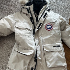 Beige dunjacka från Canada Goose - Säljer en beige dunjacka från Canada Goose med huva och pälsdetalj. Jackan har flera stora fickor, ribbade muddar och klassisk logga på bröstet. Perfekt för kalla vinterdagar och riktigt snygg till streetwear-stilen. Storlek XS men passar mig som är en L och väger 90kg 176 cm.