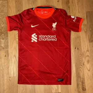 Liverpool FC röd matchtröja Nike 147-158 CM - Liverpool FCs officiella matchtröja från Nike i rött med tunna diagonala linjer. Tröjan har korta ärmar, broderat klubbmärke och sponsortryck på bröstet. Tillverkad i Dri-FIT-material som andas och håller dig sval under matchen.