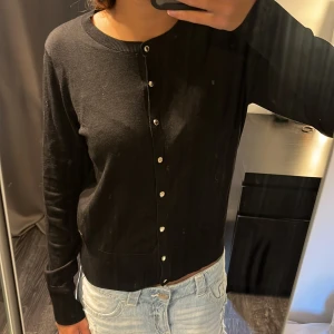 Svart kofta från Holly & Whyte, Lindex - Svart kofta från Holly & Whyte med rund halsringning och silvriga knappar framtill. Långärmad och i mjuk bomullsblandning, perfekt att slänga över en t-shirt eller linne. Enkel och stilren design som funkar till det mesta.