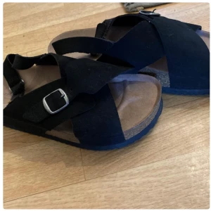 Svarta sandaler med spänne - Snygga svarta sandaler med korsade remmar i mocka och justerbart metallspänne. Fotbädden är i beige och sulan är platt och bekväm. Perfekta för varma dagar och enkel att matcha med olika outfits. Använda en gång!