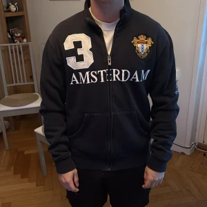 Svart zip hoodie Amsterdam Sports - Svart hoodie med dragkedja från Amsterdam Sports. Stora vita tryck med siffran 3 och texten AMSTERDAM fram och bak, broderat märke på bröstet och tryck på ärmen. Mjuk bomullskänsla och hög krage. Perfekt för en sportig och chill look.