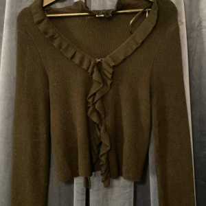 Brun volangkofta från BikBok XS - Supersöt brun kofta från BikBok i stickat material. Den har långa ärmar, volangdetaljer längs ringningen och framtill, samt en croppad passform. Perfekt att styla med jeans eller kjol för en trendig look.