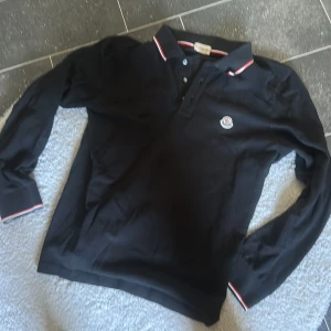 Svart långärmad pikétröja Moncler - Svart långärmad pikétröja från Moncler med klassisk krage och knäppning framtill. Tröjan har diskret Moncler-logga på bröstet och snygga rödvita detaljer på krage och ärmslut. Tillverkad i bomull för en skön och stilren känsla.