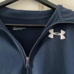 Blå zipjacka från Under Armour - Mörkblå zipjacka från Under Armour med vitt logotryck på bröstet. Jackan har lång ärm, hög krage och dragkedja hela vägen. Modellen är loose fit och passar perfekt till träning eller chill. Materialet är mjukt och stretchigt för extra komfort.