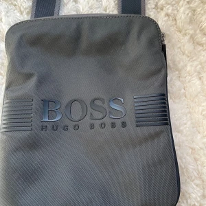 Grå axelväska från Hugo Boss - Snygg grå axelväska från Hugo Boss med justerbar rem och dragkedja upptill. Väskan har stilren design med BOSS-logga framtill och praktiskt innerfack. Perfekt storlek för mobil, plånbok och andra essentials. Materialet är slitstark polyester.