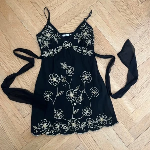 Svart blommig kortklänning med broderi - Supersöt svart miniklänning från Tfnc med tunna axelband och knytband i midjan. Klänningen har  blommiga gulddetaljer över hela framsidan och v-ringning. Skulle säga storlek S men funkar även på Xs då man kan dra åt midjan.