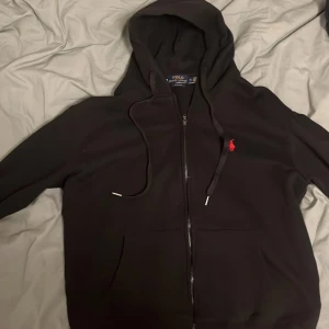 Svart hoodie från Polo Ralph Lauren - Säljer en stilren svart hoodie från Polo Ralph Lauren med dragkedja och en liten röd logga på bröstet. Perfekt för en avslappnad look. Den har en justerbar huva och långa ärmar.