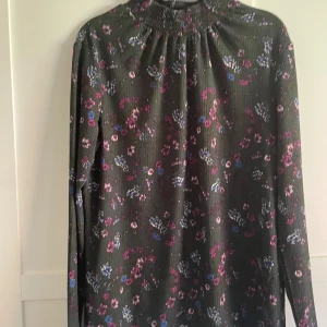 Blommig långärmad blus  - Mörkgrön långärmad blus från Jacqueline de Yong med blommigt mönster i lila, blått och rosa. Blusen har hög smockad krage och är i ett mjukt, ribbat material. Perfekt för dig som gillar en romantisk och chill stil.