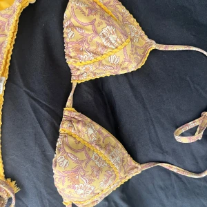 Gul och lila blommig bikini med knyt - Superfin bikini i gul och lila med blommigt mönster och söta volangdetaljer. Triangeltopp med knyt i rygg och nacke samt matchande trosa med knyt i sidorna och små tofsar. 