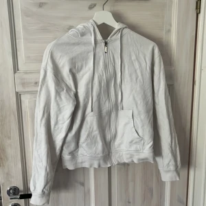 Vit hoodie med dragkedja från Cubus - Vit hoodie från Cubus med dragkedja framtill och huva med snörning. Tröjan har två stora fickor fram och är tillverkad i mjuk bomullsmix. Perfekt för chill dagar eller när du vill ha en enkel och clean look.