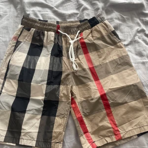 Burberry rutiga shorts - Snygga shorts med klassiskt Burberry-rutigt mönster i beige, svart, vitt och röda detaljer. De har elastisk midja med vit snörning och är tillverkade i ett lätt och svalt material, perfekt för sommaren. Passformen är normal och de har fickor på sidorna.