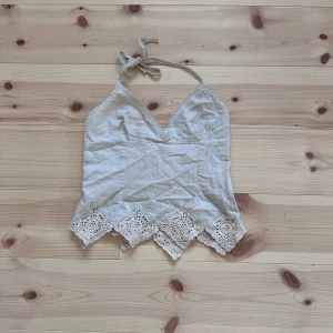 Beige linnetopp med spetsdetaljer - Supersöt  linnetopp med knyt i nacken och v-ringning. Toppen har spetskant nertill med virkade detaljer som ger en bohemisk känsla. Perfekt för varma dagar och somriga outfits.
