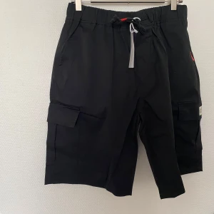 Svarta cargoshorts med snörning - Svarta cargoshorts med elastisk midja och snörning framtill. Shortsen har stora sidofickor med lock och en bakficka. Perfekta för en chill och avslappnad stil under varma dagar. Helt ny finns M-XXL kvar 