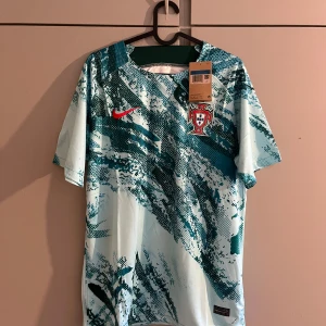 Portugal Nike fotbollströja  - Portugal fotbollströja från Nike med korta ärmar. Tröjan är vit med blått och turkost abstrakt mönster, röd Nike-logga och Portugals landslagsemblem på bröstet. Tillverkad i lätt och ventilerande material, perfekt för match eller träning.