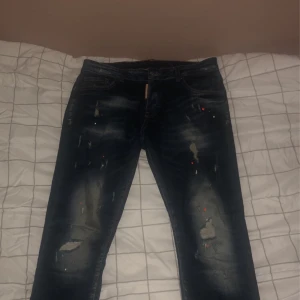 Mörkblå Dsquared2 jeans med slitningar - Säljer ett par mörkblå jeans från Dsquared2 med coola slitningar och färgstänk. Jeansen har klassisk femficksdesign, orange sömmar och en snygg patch med Rom-motiv bak. Perfekta för dig som gillar streetstyle och vill sticka ut. Äkta såklart saknas kvitto🔥