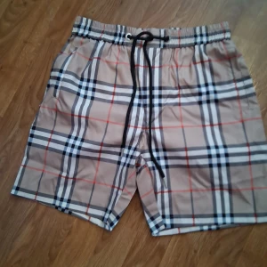 Beige rutiga shorts från Burberry - Snygga beige shorts från Burberry med klassiskt rutigt mönster i svart, vitt och rött. De har elastisk midja med svart snörning och sidofickor, samt en bakficka. Perfekta för sommaren och ger en clean, lyxig vibe.