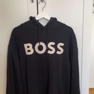 Svart hoodie från BOSS med tryck - Svart hoodie från BOSS i storlek L med stor vit BOSS-logga framtill. Klassisk huva med snörning. Skön och stilren modell, nypris 1200kr