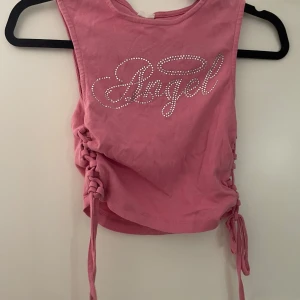 Rosa croppad topp Urban Outfitters - Säljer en rosa croppad topp från Urban Outfitters med glittrande strasstenar som bildar texten 'Angel' framtill. Toppen har snörning i sidorna för en snygg, justerbar passform och är ärmlös. Perfekt för dig som gillar statement-plagg och trendiga detaljer.