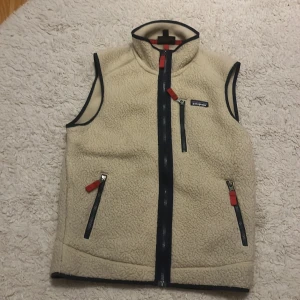 Beige fleeceväst från Patagonia S - Beige fleeceväst från Patagonia i storlek S. Västen har hög krage, tre fickor med dragkedja och snygga svarta och röda detaljer. Tillverkad i 100% återvunnen polyester och perfekt för lager-på-lager under kyliga dagar.