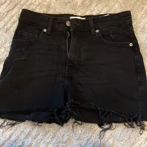 Svarta jeansshorts med fransig kant - Snygga svarta jeansshorts med hög midja och rå, fransig kant nertill. Klassisk femficksmodell med slitna detaljer på fickorna för en cool, avslappnad vibe. Perfekta för sommardagar och festivaler.