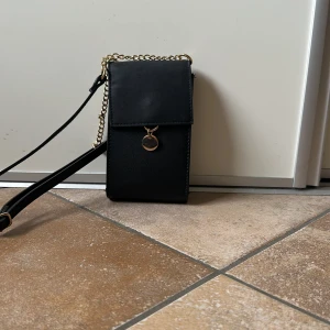 Svart axelväska med guldkedja - Snygg svart axelväska i stilren design med guldfärgad kedja och ett litet runt hänge framtill. Perfekt storlek för mobilen och det viktigaste. Väskan har ett lock och justerbar axelrem för extra bekvämlighet.