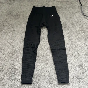 Svarta leggings från Gymshark - Säljer ett par svarta träningsleggings från Gymshark med hög midja och diskret hålmönster längs benen. De är stretchiga och har en tight passform, perfekt för träning. Gymshark-loggan syns både fram och bak i vitt. Sitter tight och snyggt.