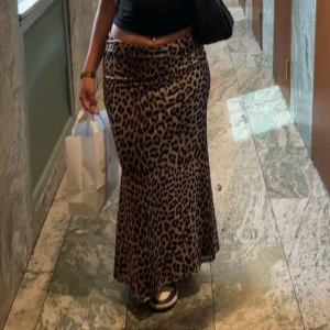 Lång leopardkjol med utsvängd passform - Säljer en långkjol med leopardmönster i bruna och svarta toner. Kjolen har en snygg utsvängd form som sitter tajt över höfterna och faller snyggt ner mot fötterna. Perfekt för dig som vill sticka ut med en trendig look.