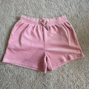 Rosa velourshorts Juicy Couture - Supermjuka rosa shorts från Juicy Couture i velour med snörning i midjan och en bakficka. Klassisk broderad JC-logga framtill. Perfekta för chill dagar eller när du vill ha en söt och bekväm look.