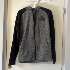Grå och svart hoodie från Adidas S - Säljer en gråmelerad hoodie från Adidas med svarta ärmar och svart logga på bröstet. Tröjan har hel dragkedja, huva med snörning och två fickor framtill. Insidan är mjuk och fleecefodrad, perfekt för kyliga dagar.