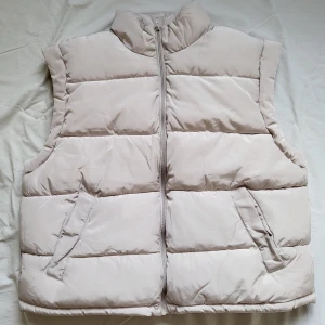 Beige dunväst från H&M XL - Snygg beige dunväst från H&M i storlek XL. Västen har quiltad design, hög krage och dragkedja framtill. Perfekt att slänga över en hoodie för en chill look. Två fickor framtill och lätt vadderad för extra värme. Boxy fit.