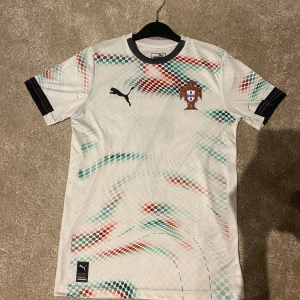 Portugal Ronaldo 7 fotbollströja Puma - Portugal landslagströja från Puma med Ronaldo 7 på ryggen. Vit bas med prickigt mönster i rött, blått och grönt. Kortärmad modell med svarta detaljer vid ärmslut och krage. Tillverkad i lätt och ventilerande funktionsmaterial.