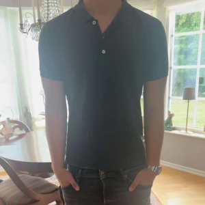 Oscar Jacobsson pike - Klassisk svart pikétröja från Oscar Jacobson i storlek S. Tröjan har korta ärmar, krage och knäppning med två knappar framtill. Tillverkad i bomull för en skön och stilren känsla. Diskret broderad logga nertill på sidan.