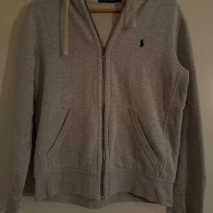 Grå hoodie från Polo Ralph Lauren - Mysig grå hoodie från Polo Ralph Lauren med dragkedja framtill och två fickor. Klassisk logga broderad på bröstet och justerbar huva med snören. Perfekt för en avslappnad stil. PRIS KAN DISKUTERAS!